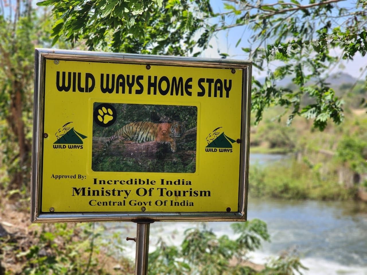 Yellow Wild Ways Home Stay signboard with a tiger photograph, beside a river